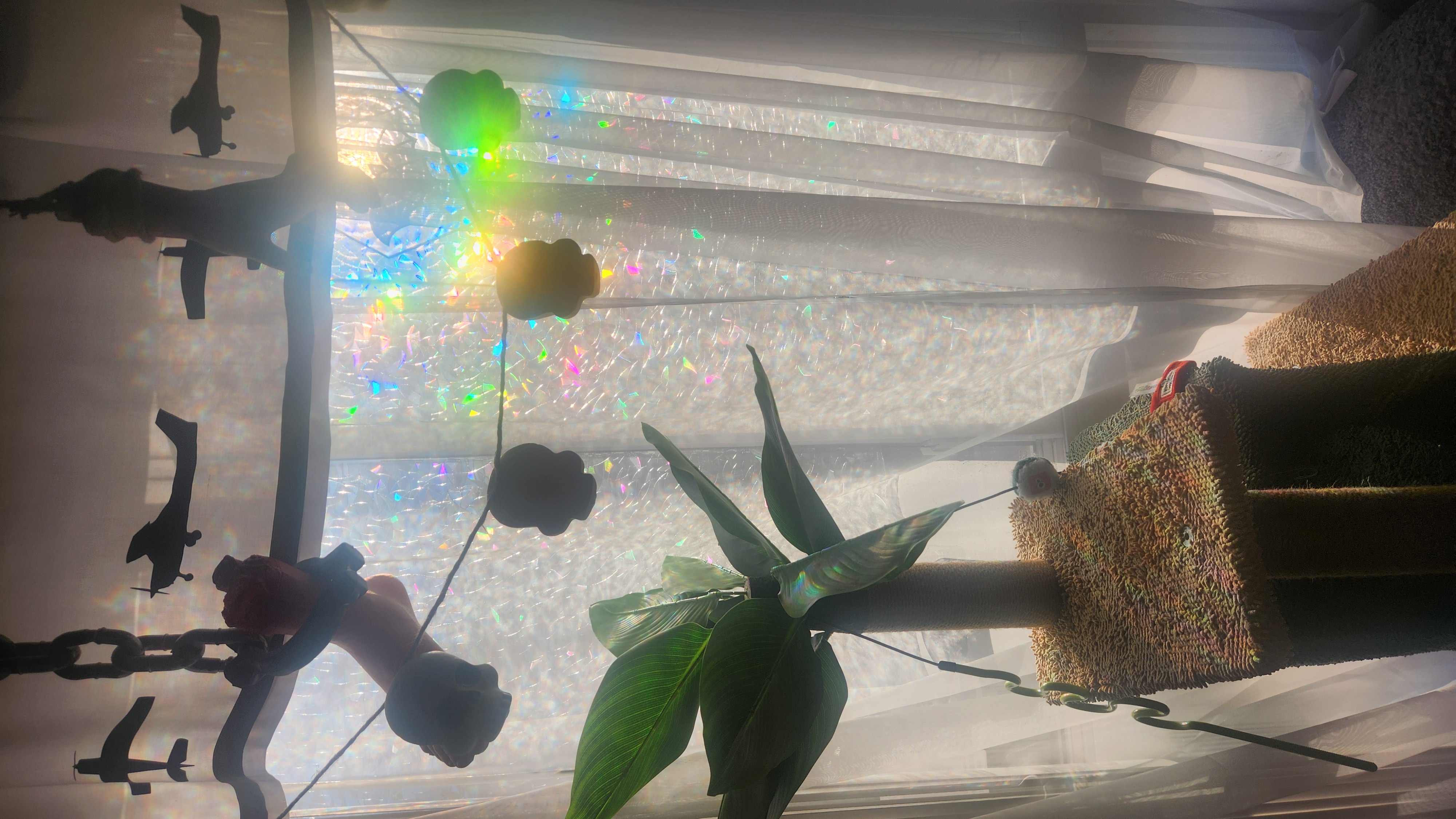 a cat tree with a fake plant on top sits in front of the morning window with overcast sunlight filtering through a prism sticker to make a rainbow reflection