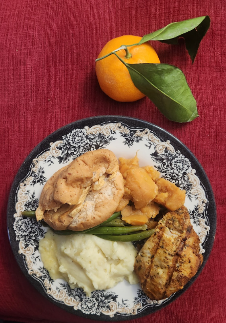 a plate with chicken, green beans, and two different kinds of potatoes sits next to a mandarin orange on top of a red background