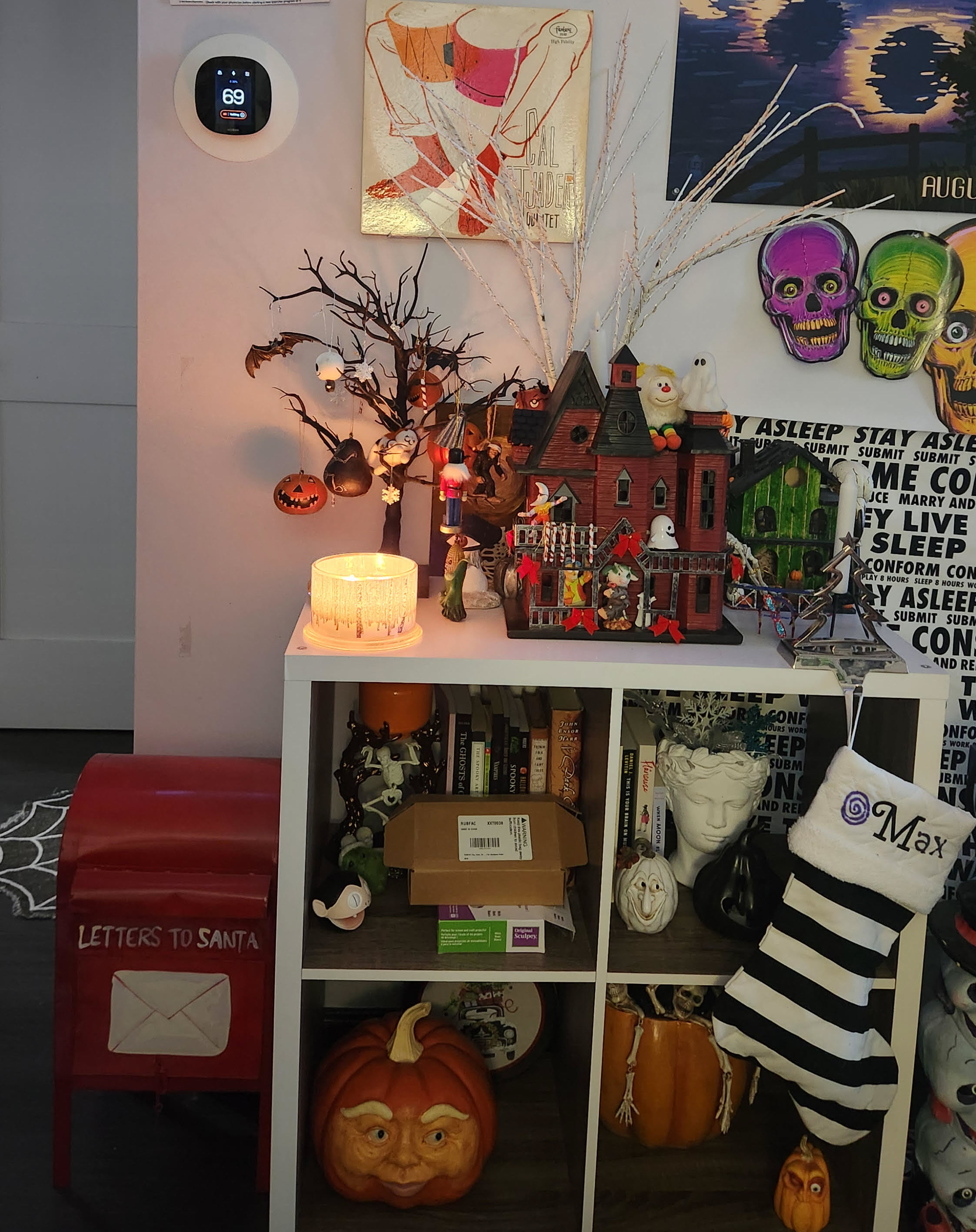 A white cubed shelf with a red and black gothic house on top. The house is decorated for Yule with red bows and candy canes, with a white branched tree behind it. Beside it is a black tabletop creepy tree with various ornaments.